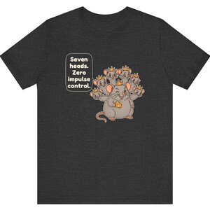 May include: A dark gray t-shirt featuring a graphic of a seven-headed mouse wearing small gold crowns, holding a piece of yellow cheese, and a speech bubble with the text: Seven heads. Zero impulse control.