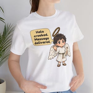 May include: A white t-shirt featuring a graphic of a cartoon angel with brown hair, a crooked halo, and white wings. The angel holds a scroll and waves. A yellow speech bubble contains the text: Halo crooked. Message delivered.