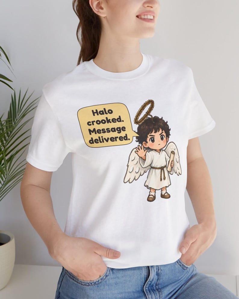 May include: A white t-shirt featuring a graphic of a cartoon angel with brown hair, a crooked halo, and white wings. The angel holds a scroll and waves. A yellow speech bubble contains the text: Halo crooked. Message delivered.