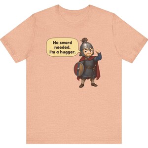 May include: A peach-colored t-shirt featuring a cartoon illustration of a warrior in chainmail and a helmet with a small animal on top. A speech bubble contains the text: No sword needed. I'm a hugger. The warrior holds a round shield.