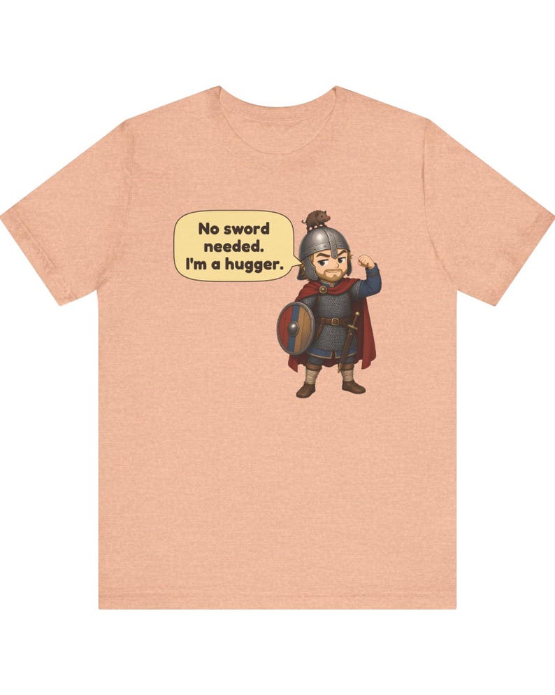 May include: A peach-colored t-shirt featuring a cartoon illustration of a warrior in chainmail and a helmet with a small animal on top. A speech bubble contains the text: No sword needed. I'm a hugger. The warrior holds a round shield.