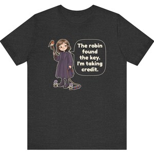 May include: A dark heather gray short-sleeve t-shirt featuring a cartoon illustration of a person in a long coat holding a key with a robin perched on it. A speech bubble contains the text: The robin found the key. I'm taking credit.
