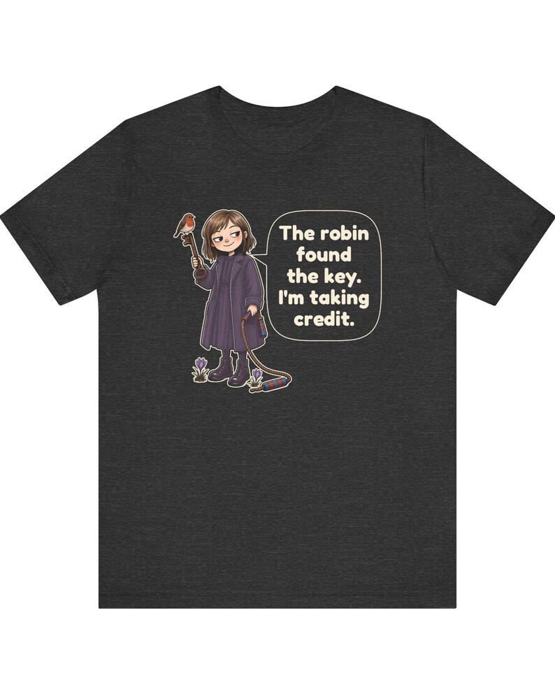 May include: A dark heather gray short-sleeve t-shirt featuring a cartoon illustration of a person in a long coat holding a key with a robin perched on it. A speech bubble contains the text: The robin found the key. I'm taking credit.