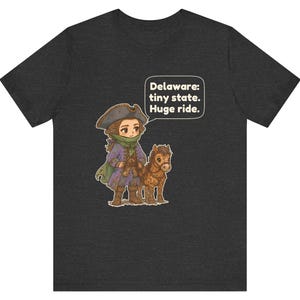 May include: A dark heather gray t-shirt featuring a cartoon illustration of a person in a tricorn hat and purple coat standing next to a small brown horse. A speech bubble contains the text: Delaware: tiny state. Huge ride.