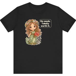 May include: A black t-shirt featuring a cartoon illustration of a character with long brown hair, a flower crown, and a green dress holding a pomegranate. A speech bubble next to the character contains the text: Six seeds. Totally worth it.