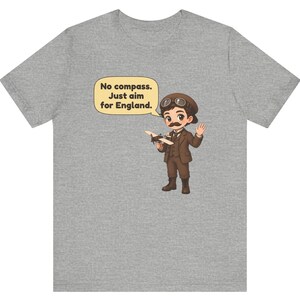 May include: A heather gray t-shirt featuring a cartoon illustration of a pilot with a mustache, wearing a brown suit and aviator cap, holding a small airplane. A speech bubble contains the text: No compass. Just aim for England.
