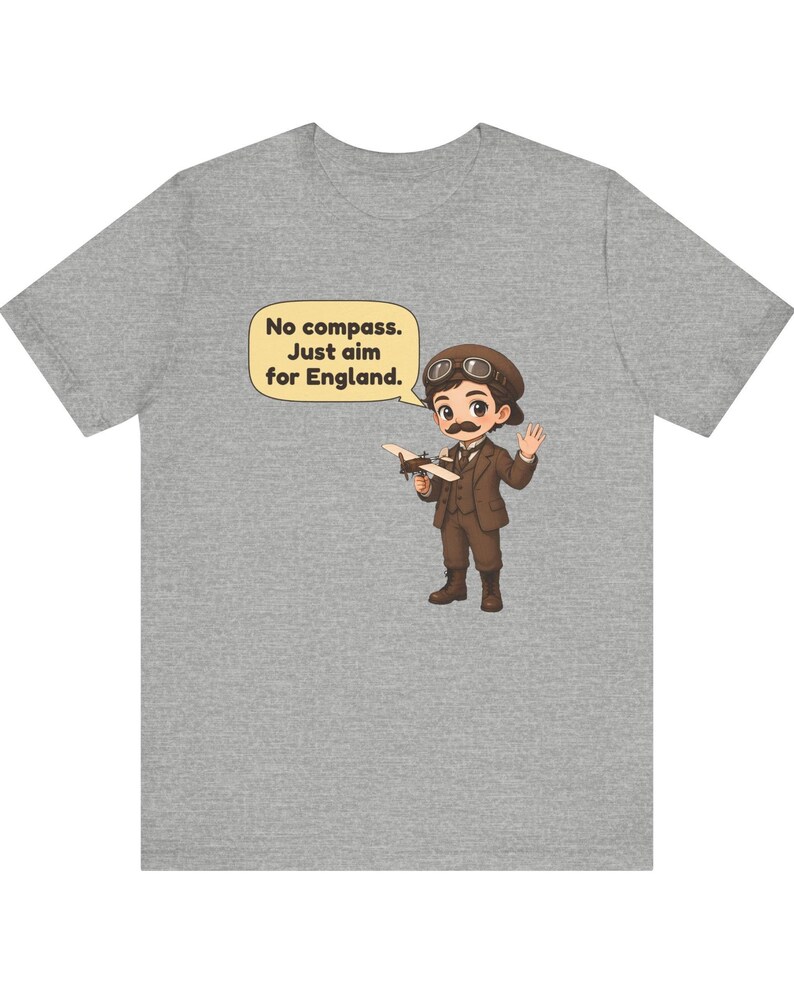 May include: A heather gray t-shirt featuring a cartoon illustration of a pilot with a mustache, wearing a brown suit and aviator cap, holding a small airplane. A speech bubble contains the text: No compass. Just aim for England.