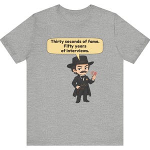 Pu&ograve; includere: Una t-shirt grigio melange con un'illustrazione a cartone animato di un uomo in abito nero, cappello e baffi che tiene in mano delle carte da gioco. Un fumetto sopra la figura contiene il testo: Thirty seconds of fame. Fifty years of interviews.