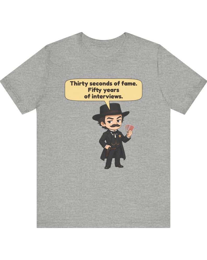 Pu&ograve; includere: Una t-shirt grigio melange con un'illustrazione a cartone animato di un uomo in abito nero, cappello e baffi che tiene in mano delle carte da gioco. Un fumetto sopra la figura contiene il testo: Thirty seconds of fame. Fifty years of interviews.