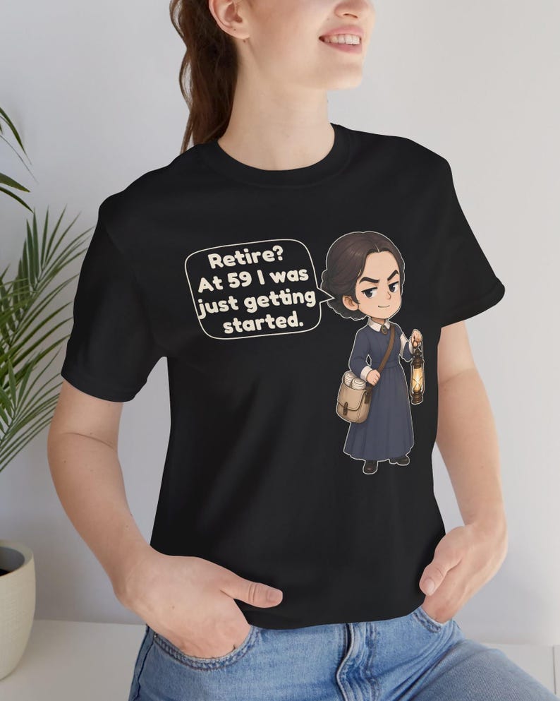May include: A black t-shirt featuring a cartoon illustration of a person in a dark blue dress carrying a tan satchel and a lit lantern. A speech bubble next to the character contains the text: Retire? At 59 I was just getting started.