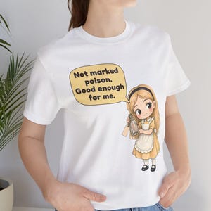 May include: A white t-shirt featuring a graphic of a blonde character in a yellow dress and white apron holding a glass bottle. A speech bubble above the character contains the text: Not marked poison. Good enough for me.
