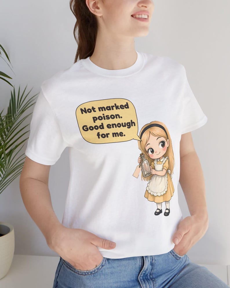 May include: A white t-shirt featuring a graphic of a blonde character in a yellow dress and white apron holding a glass bottle. A speech bubble above the character contains the text: Not marked poison. Good enough for me.