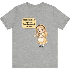 May include: A heather gray t-shirt featuring a cartoon illustration of a blonde character in a yellow dress and white apron holding a glass bottle. A speech bubble above the character contains the text: Not marked poison. Good enough for me.
