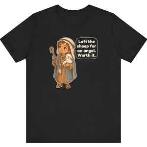 May include: A black t-shirt featuring a cartoon illustration of a shepherd holding a small white sheep and a wooden staff. The shepherd wears a brown tunic and a blue cloak. A speech bubble contains the text: Left the sheep for an angel. Worth it.