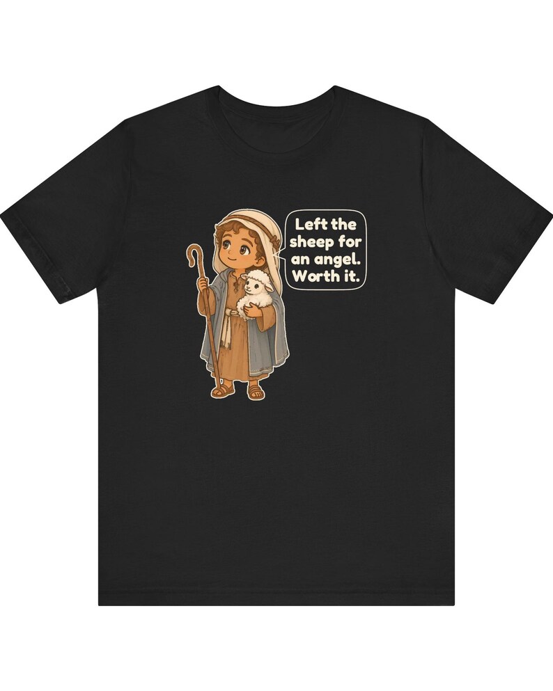 May include: A black t-shirt featuring a cartoon illustration of a shepherd holding a small white sheep and a wooden staff. The shepherd wears a brown tunic and a blue cloak. A speech bubble contains the text: Left the sheep for an angel. Worth it.