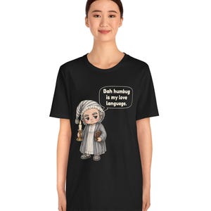 Puede incluir: Una camiseta negra de manga corta con una ilustraci&oacute;n de dibujos animados de un personaje con gorro de dormir y bata que sostiene una vela y un libro. Un globo de texto sobre el personaje contiene el texto, "Bah humbug is my love language."