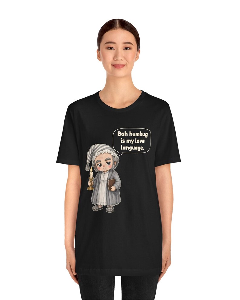 Puede incluir: Una camiseta negra de manga corta con una ilustraci&oacute;n de dibujos animados de un personaje con gorro de dormir y bata que sostiene una vela y un libro. Un globo de texto sobre el personaje contiene el texto, "Bah humbug is my love language."