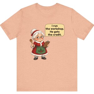 K&ouml;nnte beinhalten: Ein pfirsichfarbenes T-Shirt mit einer Cartoon-Illustration einer Frau mit wei&szlig;em Haar in einem rot-wei&szlig;en Outfit im Weihnachtsmann-Stil, die einen Holzl&ouml;ffel und ein Klemmbrett h&auml;lt, mit einer Sprechblase: I run the workshop. He gets the credit.