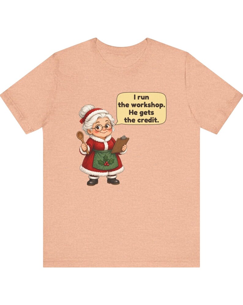 K&ouml;nnte beinhalten: Ein pfirsichfarbenes T-Shirt mit einer Cartoon-Illustration einer Frau mit wei&szlig;em Haar in einem rot-wei&szlig;en Outfit im Weihnachtsmann-Stil, die einen Holzl&ouml;ffel und ein Klemmbrett h&auml;lt, mit einer Sprechblase: I run the workshop. He gets the credit.