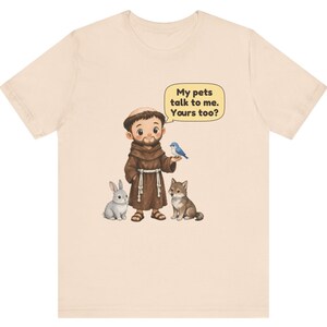 May include: A light beige t-shirt featuring a cartoon illustration of a monk in a brown robe standing with a grey rabbit, a brown wolf, and a blue bird. A yellow speech bubble above the monk contains the text: My pets talk to me. Yours too?