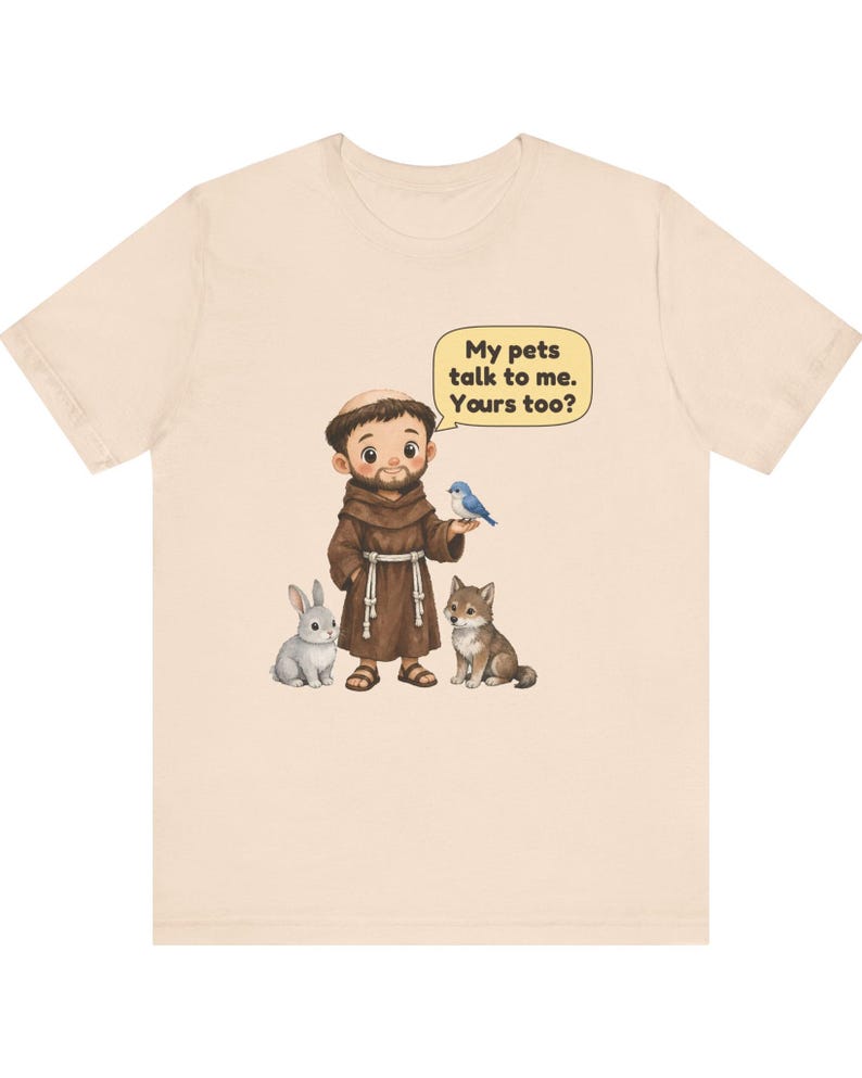 May include: A light beige t-shirt featuring a cartoon illustration of a monk in a brown robe standing with a grey rabbit, a brown wolf, and a blue bird. A yellow speech bubble above the monk contains the text: My pets talk to me. Yours too?