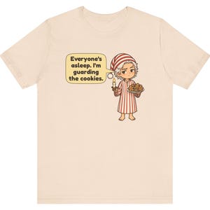 Op de afbeelding: Een lichtbeige t-shirt met een cartoonillustratie van een persoon in een rood-wit gestreepte pyjama en een bijpassende slaapmuts. De persoon houdt een brandende kaars en een bord met koekjes vast, met een tekstballon: Everyone's asleep. I'm guarding the cookies.