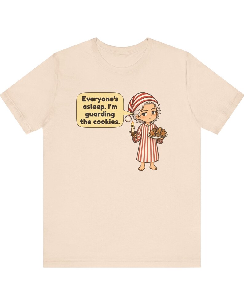 Op de afbeelding: Een lichtbeige t-shirt met een cartoonillustratie van een persoon in een rood-wit gestreepte pyjama en een bijpassende slaapmuts. De persoon houdt een brandende kaars en een bord met koekjes vast, met een tekstballon: Everyone's asleep. I'm guarding the cookies.