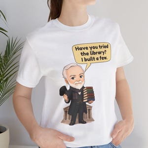 May include: A white t-shirt featuring a cartoon illustration of a bearded man in a dark coat and top hat holding a stack of books. A speech bubble above him contains the text: Have you tried the library? I built a few.