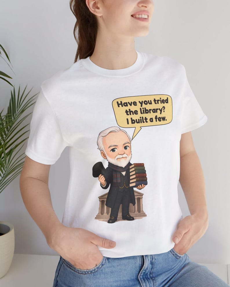 May include: A white t-shirt featuring a cartoon illustration of a bearded man in a dark coat and top hat holding a stack of books. A speech bubble above him contains the text: Have you tried the library? I built a few.