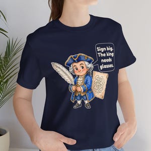 May include: A navy blue t-shirt featuring a cartoon illustration of a historical figure in a blue coat and tricorn hat holding a white quill. Next to the figure is a speech bubble with the text: Sign big. The king needs glasses.