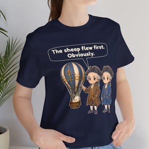 May include: A navy blue t-shirt featuring a graphic of a hot air balloon with a sheep in the basket, two figures in 18th-century attire, and a speech bubble containing the text: The sheep flew first. Obviously.
