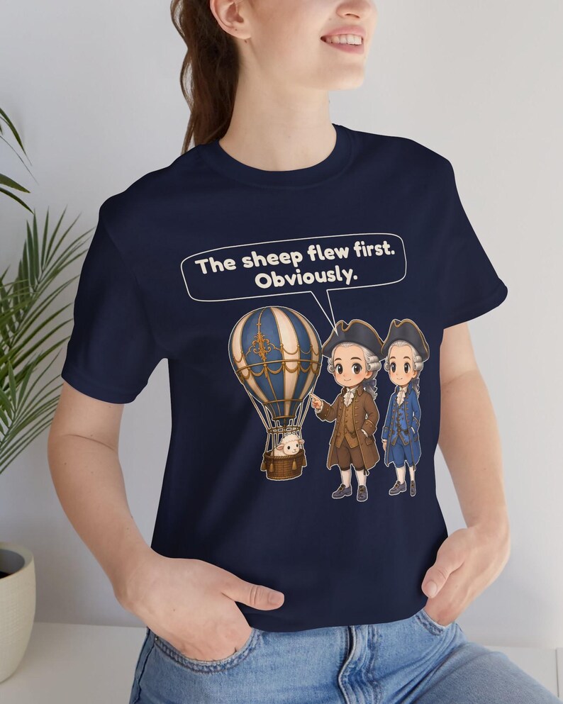 May include: A navy blue t-shirt featuring a graphic of a hot air balloon with a sheep in the basket, two figures in 18th-century attire, and a speech bubble containing the text: The sheep flew first. Obviously.