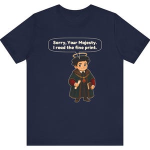 May include: A navy blue t-shirt featuring a cartoon illustration of a person in historical attire with a halo, holding a scroll and a book. A speech bubble above the character contains the text: Sorry, Your Majesty. I read the fine print.