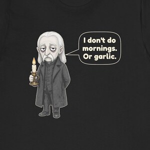 May include: A black t-shirt featuring a cartoon illustration of a pale vampire with long white hair, wearing a dark coat and holding a lit candle. A speech bubble next to the character contains the text: I don't do mornings. Or garlic.