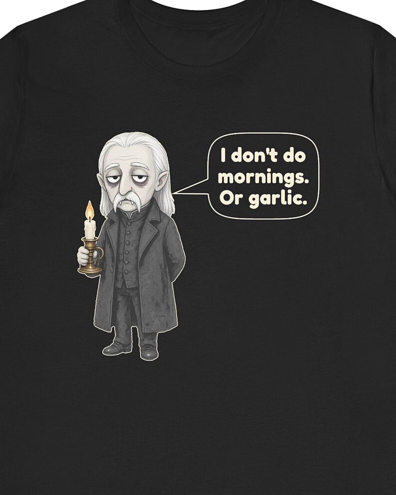 May include: A black t-shirt featuring a cartoon illustration of a pale vampire with long white hair, wearing a dark coat and holding a lit candle. A speech bubble next to the character contains the text: I don't do mornings. Or garlic.