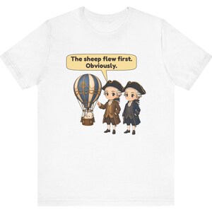 May include: A white t-shirt featuring a graphic of two figures in 18th-century attire standing next to a hot air balloon with a sheep in the basket. A speech bubble above them contains the text: The sheep flew first. Obviously.
