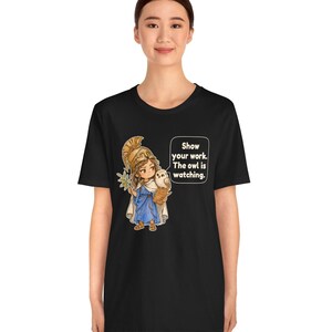 Puede incluir: Una camiseta negra de manga corta con un gr&aacute;fico de un personaje con casco dorado y t&uacute;nica azul, sosteniendo una rama de olivo y un peque&ntilde;o b&uacute;ho. Un bocadillo de texto contiene las palabras: Show your work. The owl is watching.