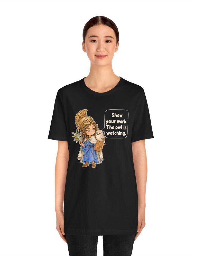 Puede incluir: Una camiseta negra de manga corta con un gr&aacute;fico de un personaje con casco dorado y t&uacute;nica azul, sosteniendo una rama de olivo y un peque&ntilde;o b&uacute;ho. Un bocadillo de texto contiene las palabras: Show your work. The owl is watching.