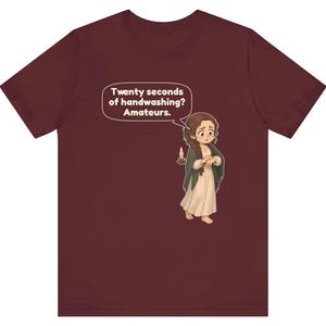 Op de afbeelding: Een bordeauxrood t-shirt met korte mouwen en een cartoonillustratie van een persoon met een lange vlecht die een kaars vasthoudt. Een tekstballon boven het personage bevat de tekst: Twenty seconds of handwashing? Amateurs.
