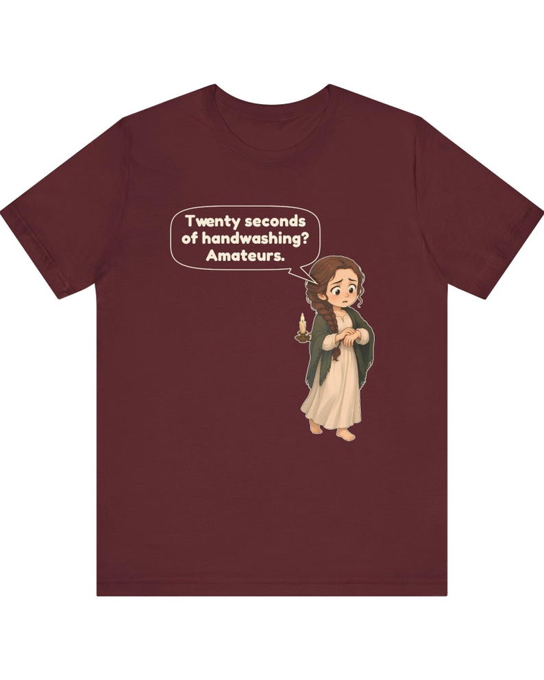 Op de afbeelding: Een bordeauxrood t-shirt met korte mouwen en een cartoonillustratie van een persoon met een lange vlecht die een kaars vasthoudt. Een tekstballon boven het personage bevat de tekst: Twenty seconds of handwashing? Amateurs.