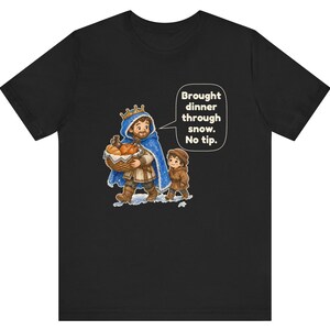May include: A black t-shirt featuring a cartoon illustration of a person wearing a blue cloak and gold crown, carrying a basket of bread, walking with a child in a brown coat. A speech bubble contains the text: Brought dinner through snow. No tip.