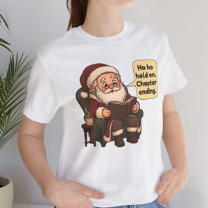 Op de afbeelding: Een wit t-shirt met een afbeelding van de Kerstman die in een groene fauteuil zit, een boek leest en een dampende mok vasthoudt. Een gele tekstballon ernaast bevat de tekst: Ho ho hold on. Chapter ending.