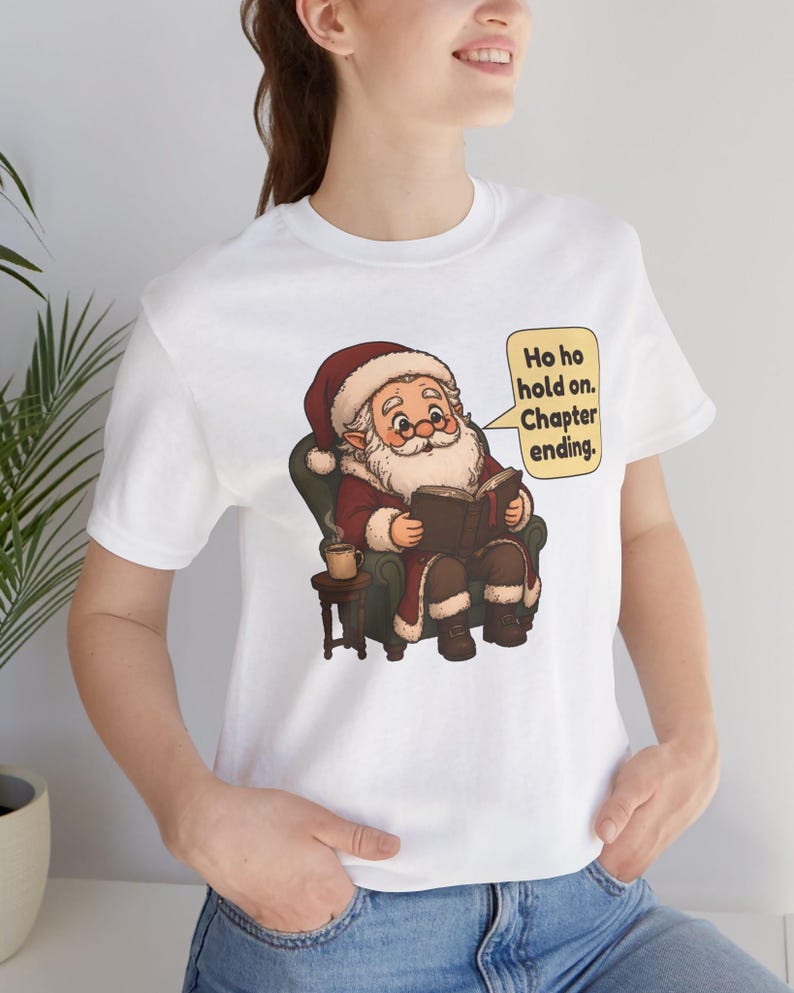 Op de afbeelding: Een wit t-shirt met een afbeelding van de Kerstman die in een groene fauteuil zit, een boek leest en een dampende mok vasthoudt. Een gele tekstballon ernaast bevat de tekst: Ho ho hold on. Chapter ending.