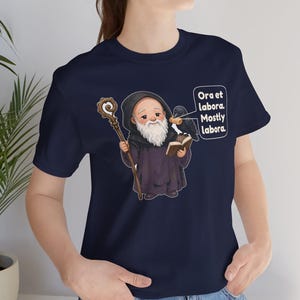 May include: A navy blue t-shirt featuring a graphic of a bearded monk in a dark robe holding a wooden staff and an open book. A black raven sits on the book, and a speech bubble contains the text: Ora et labora. Mostly labora.