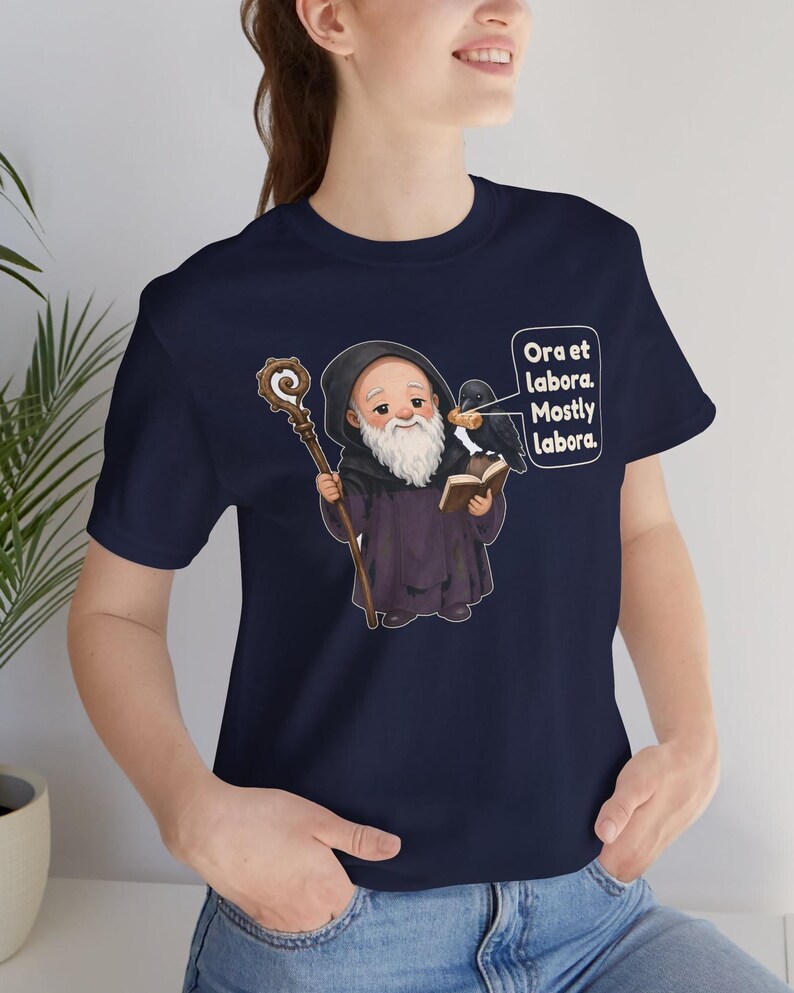 May include: A navy blue t-shirt featuring a graphic of a bearded monk in a dark robe holding a wooden staff and an open book. A black raven sits on the book, and a speech bubble contains the text: Ora et labora. Mostly labora.