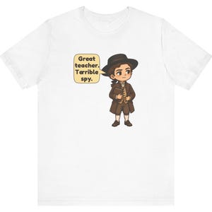 May include: A white short-sleeve t-shirt featuring a cartoon illustration of a person in 18th-century attire, including a brown coat, vest, and hat. A speech bubble next to the character contains the text: Great teacher. Terrible spy.