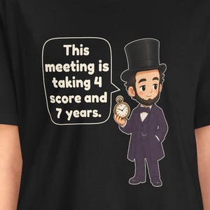 May include: A black t-shirt featuring a cartoon illustration of Abraham Lincoln wearing a top hat and suit, holding a pocket watch. A speech bubble next to him contains the text: This meeting is taking 4 score and 7 years.