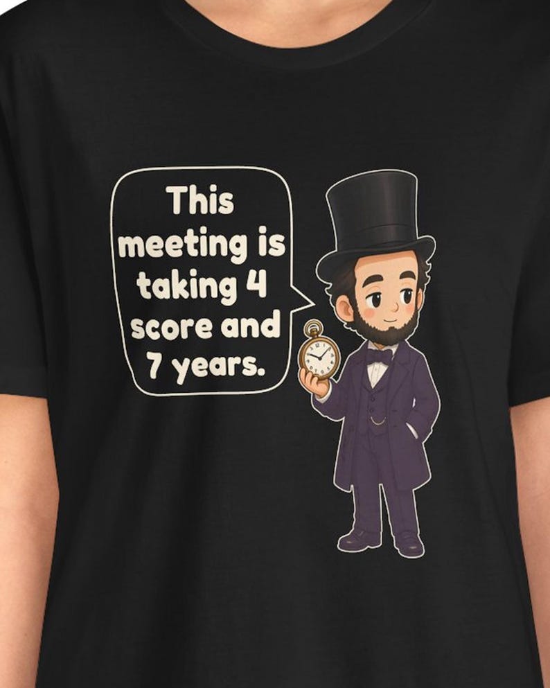 May include: A black t-shirt featuring a cartoon illustration of Abraham Lincoln wearing a top hat and suit, holding a pocket watch. A speech bubble next to him contains the text: This meeting is taking 4 score and 7 years.