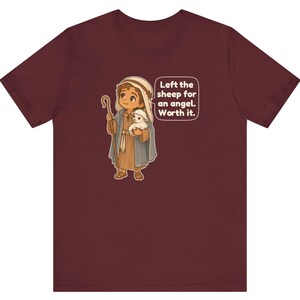 May include: A maroon short-sleeve t-shirt featuring a cartoon illustration of a shepherd holding a small white sheep and a wooden staff. A speech bubble next to the character contains the text: Left the sheep for an angel. Worth it.