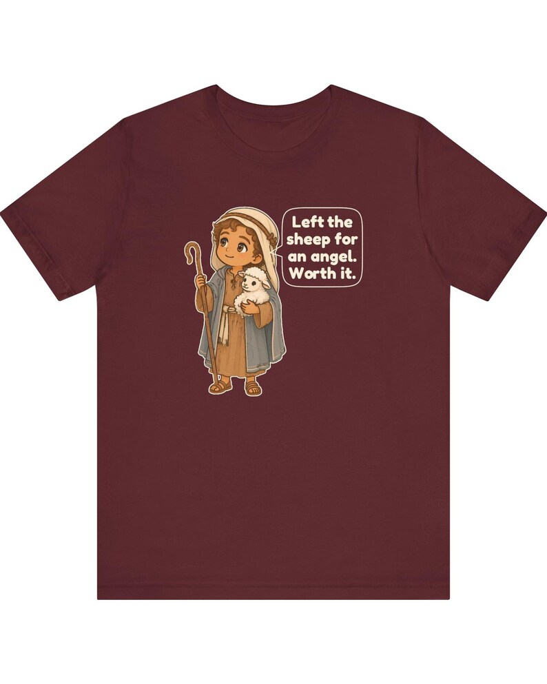 May include: A maroon short-sleeve t-shirt featuring a cartoon illustration of a shepherd holding a small white sheep and a wooden staff. A speech bubble next to the character contains the text: Left the sheep for an angel. Worth it.